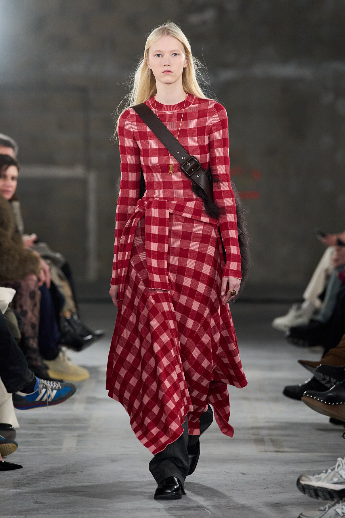 Model walking a fashion runway in a red plaid long-sleeve dress with a black crossbody belt bag and black boots.