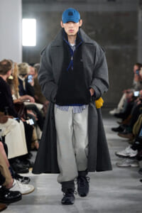 Male model on a runway wearing an oversized gray coat over a black zip-neck top, striped shirt, and baggy gray sweatpants with a blue cap and yellow gloves.