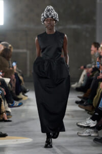 Model walking a fashion show wearing a sleeveless black dress with a high waist and a large gathered skirt; black-and-white knitted hat adds volume.