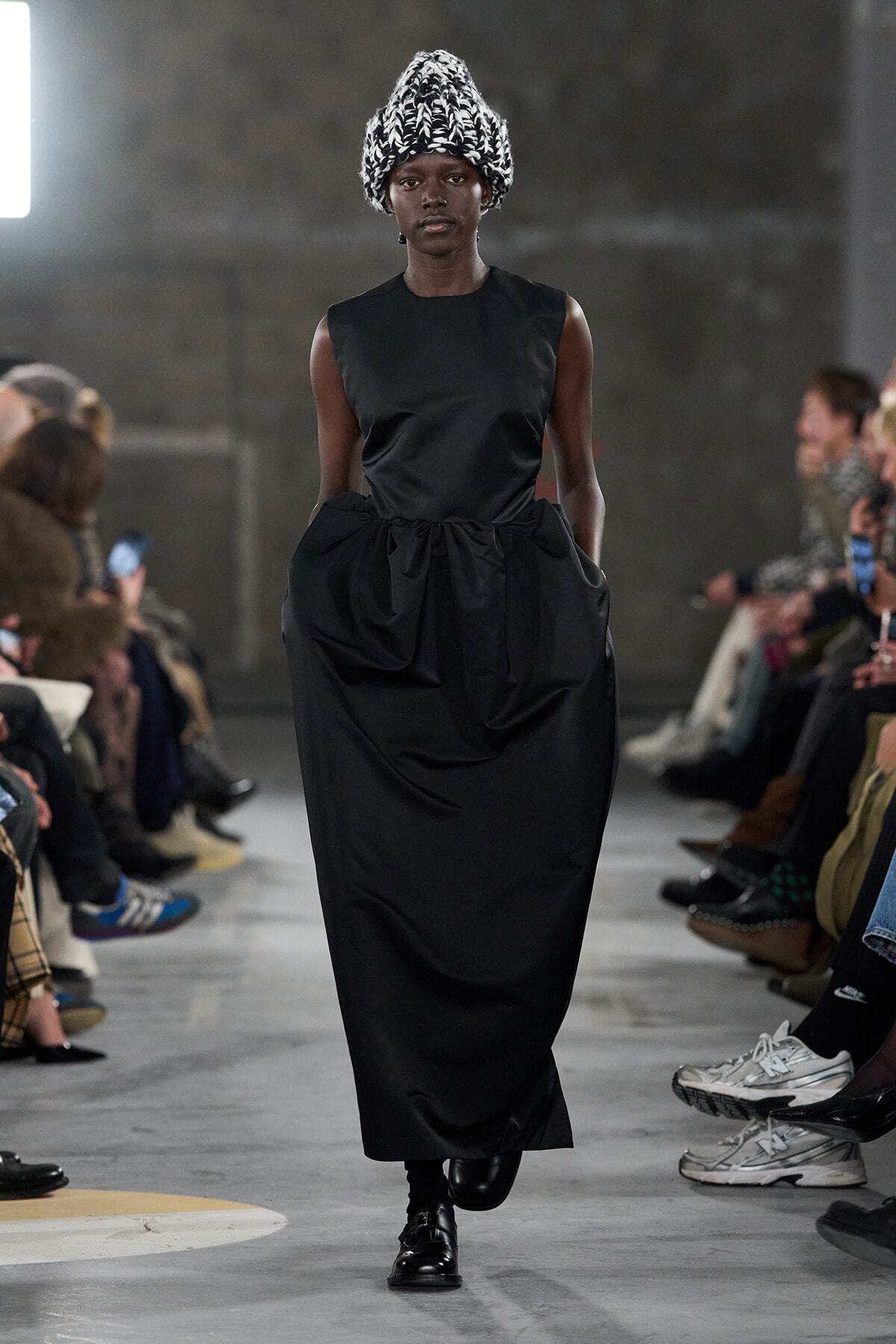 Model walking a fashion show wearing a sleeveless black dress with a high waist and a large gathered skirt; black-and-white knitted hat adds volume.