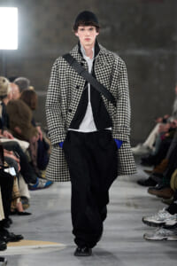 Male model on a fashion runway wearing a black-and-white houndstooth overcoat, white shirt, black pants, and a diagonal crossbody strap.