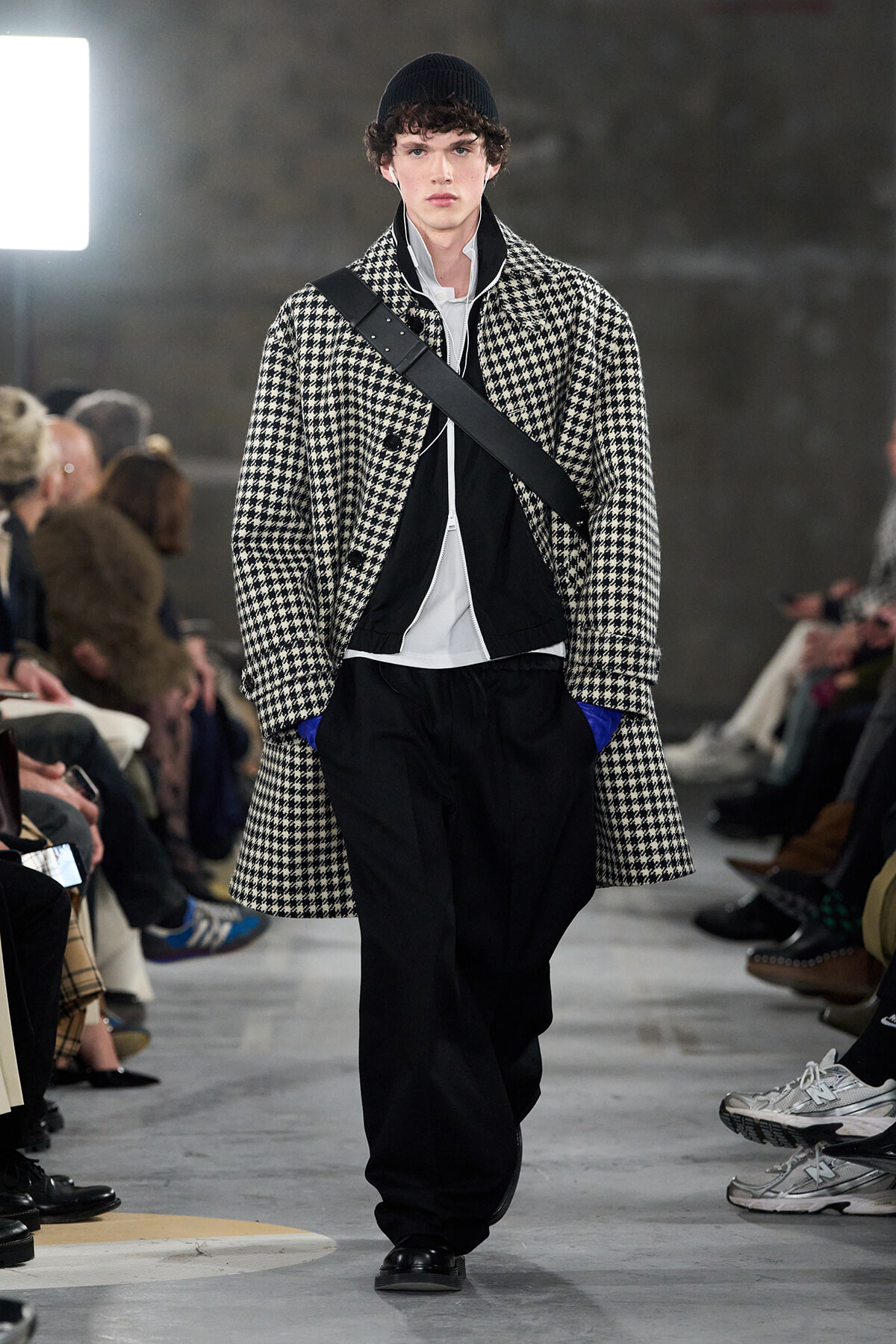 Male model on a fashion runway wearing a black-and-white houndstooth overcoat, white shirt, black pants, and a diagonal crossbody strap.