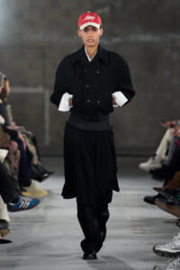 Model walking a runway in a black layered outfit with a red cap and white cuffs showing, hands in pockets.