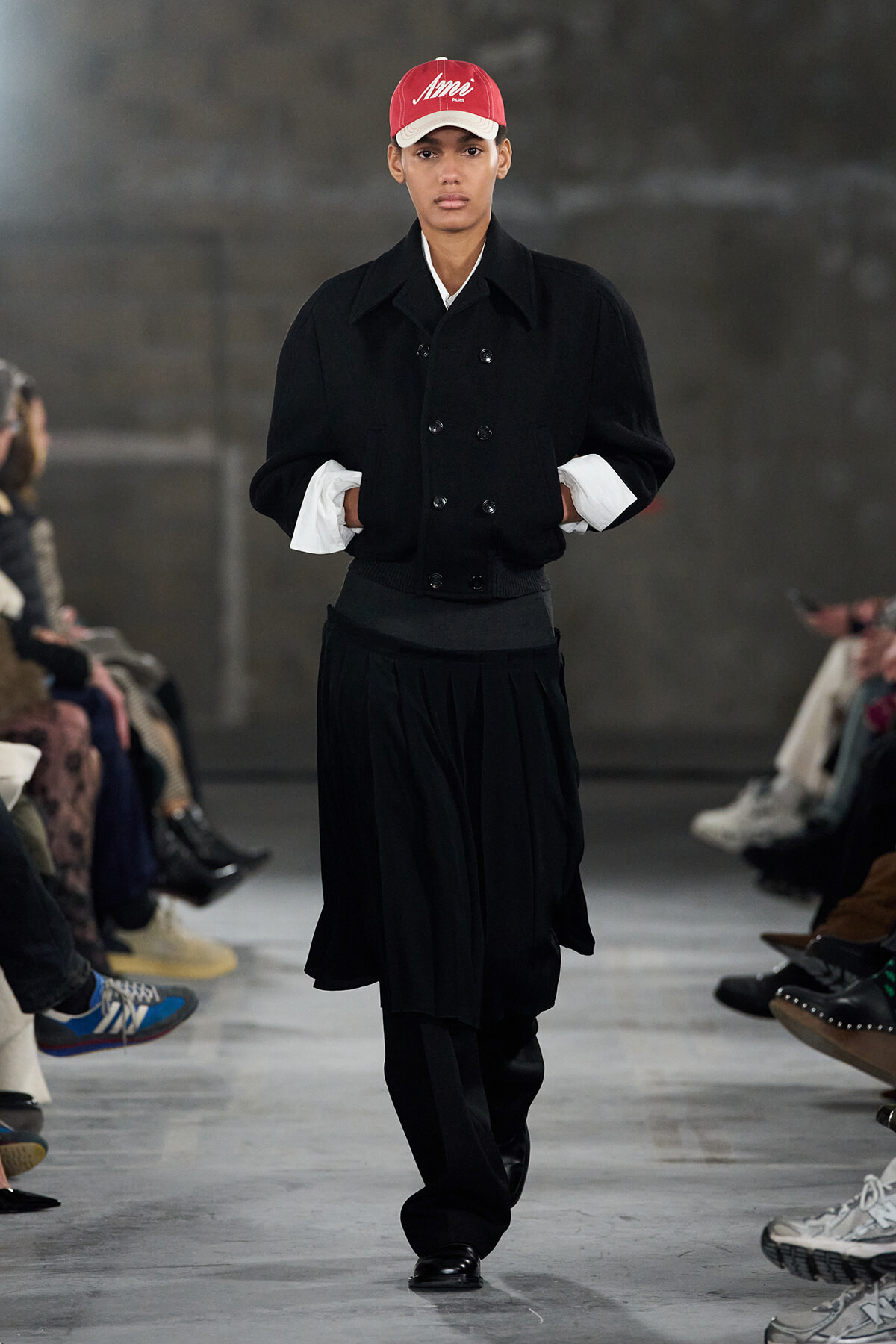 Model walking a runway in a black layered outfit with a red cap and white cuffs showing, hands in pockets.