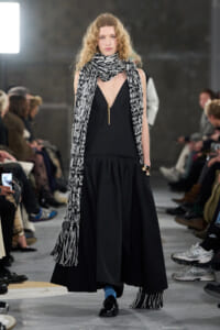 Runway model in a black V-neck dress with a long black-and-white scarf, blue socks, and black loafers.