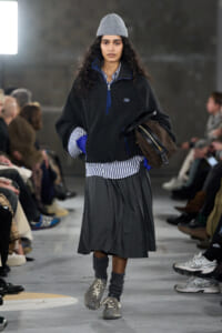 Runway model walking toward the camera in a gray knit beanie, oversized black fleece, blue-striped shirt, a pleated gray skirt, gray socks, sneakers, and a brown handbag held under her arm.