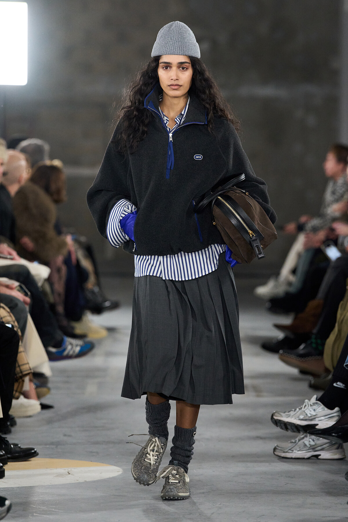 Runway model walking toward the camera in a gray knit beanie, oversized black fleece, blue-striped shirt, a pleated gray skirt, gray socks, sneakers, and a brown handbag held under her arm.