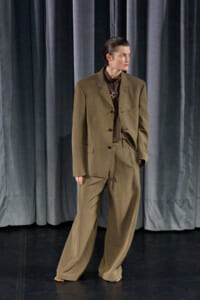 Person wearing an oversized tan suit on a dark stage with gray curtains, looking to the side and hands in pockets.