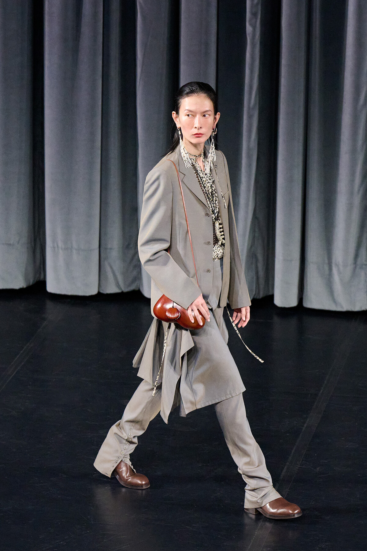 Model walking on a stage in a light gray layered suit, brown shoes, and a small brown crossbody bag against dark curtains.