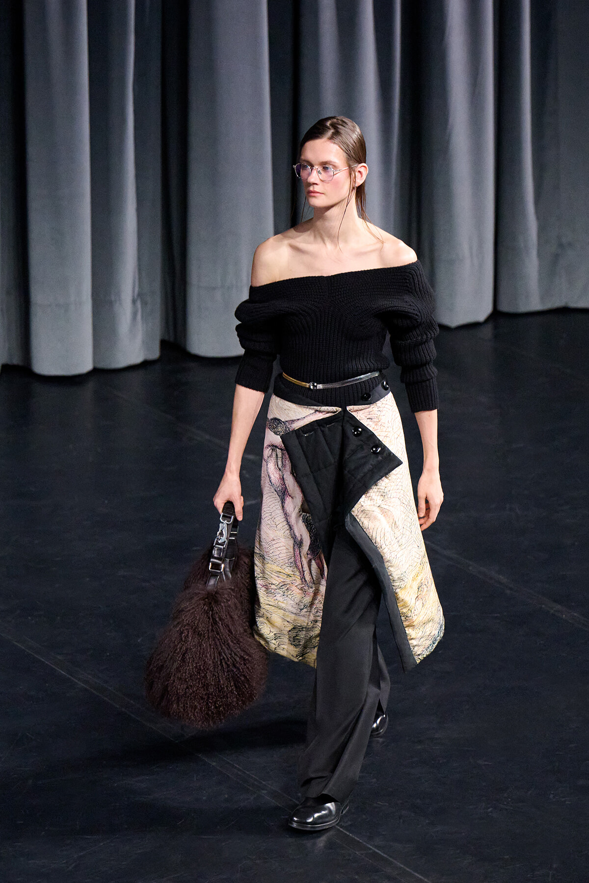 Model on a runway in an off-shoulder black knit top, layered patterned skirt over pants, and a large dark furry handbag.