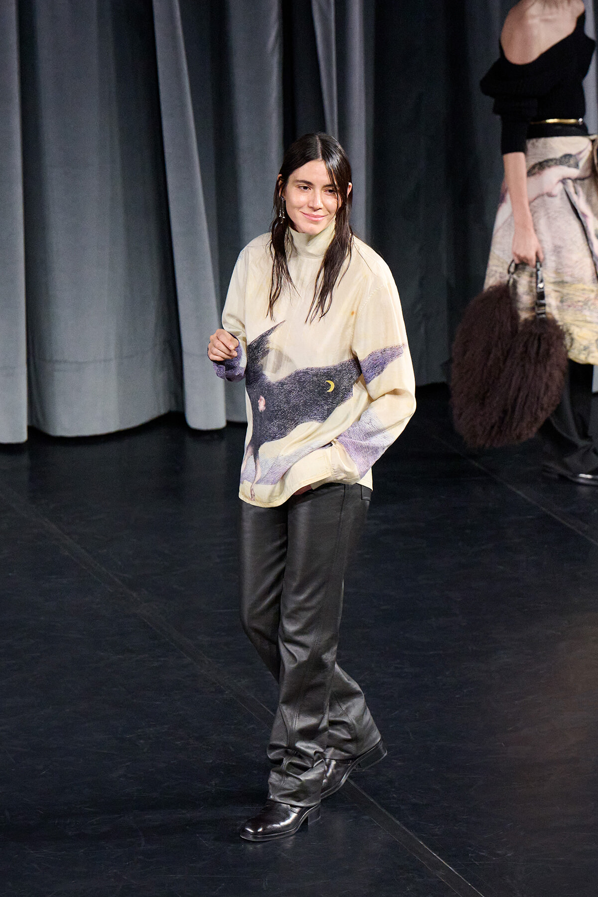 Model walking on a runway in a beige oversized sweater with a wolf print and black leather pants, backstage stage curtains in the background