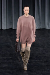 Model walking on a runway in a oversized dusty pink sweater dress with knee-high taupe boots and beige socks, carrying a small dark handbag.