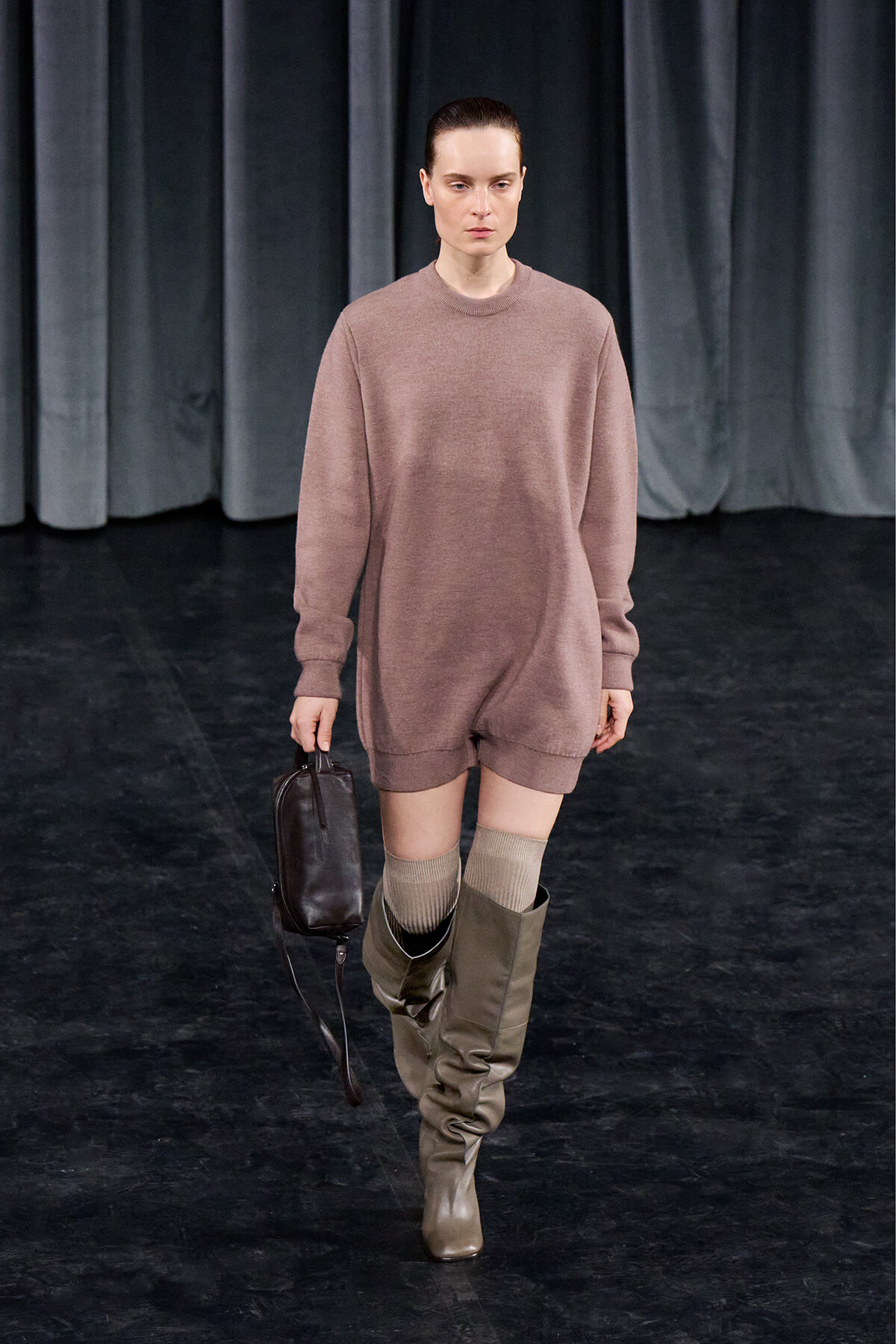 Model walking on a runway in a oversized dusty pink sweater dress with knee-high taupe boots and beige socks, carrying a small dark handbag.
