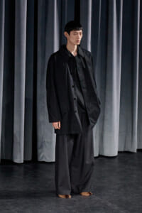 Person standing on a dark stage wearing an oversized black coat and wide-leg black trousers, with dark gray curtains in the background.
