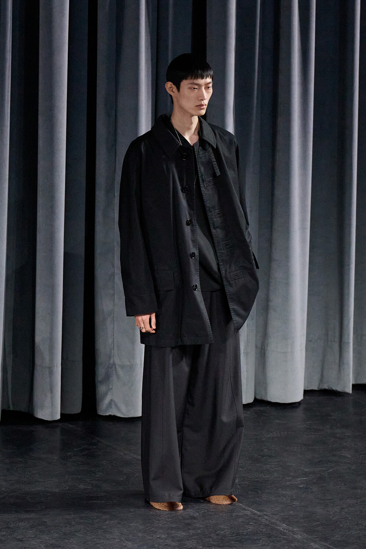 Person standing on a dark stage wearing an oversized black coat and wide-leg black trousers, with dark gray curtains in the background.