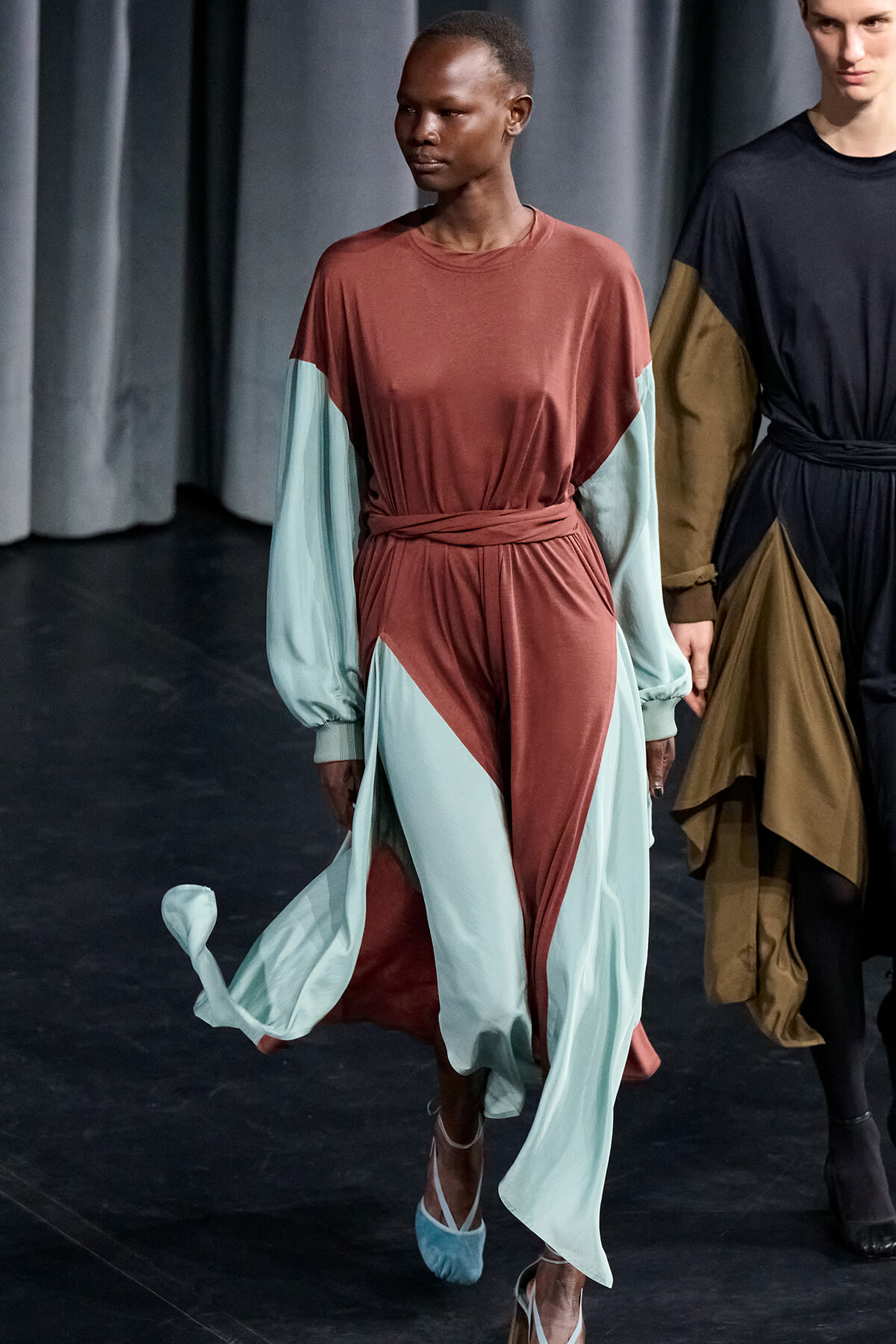 Runway model wearing a color-block dress with rust bodice, pale teal sleeves and panels, and a wrapped waist with flowing skirt.