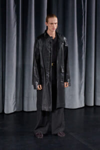 Male model wearing a long black raincoat on a dark stage with gray curtains in the background.