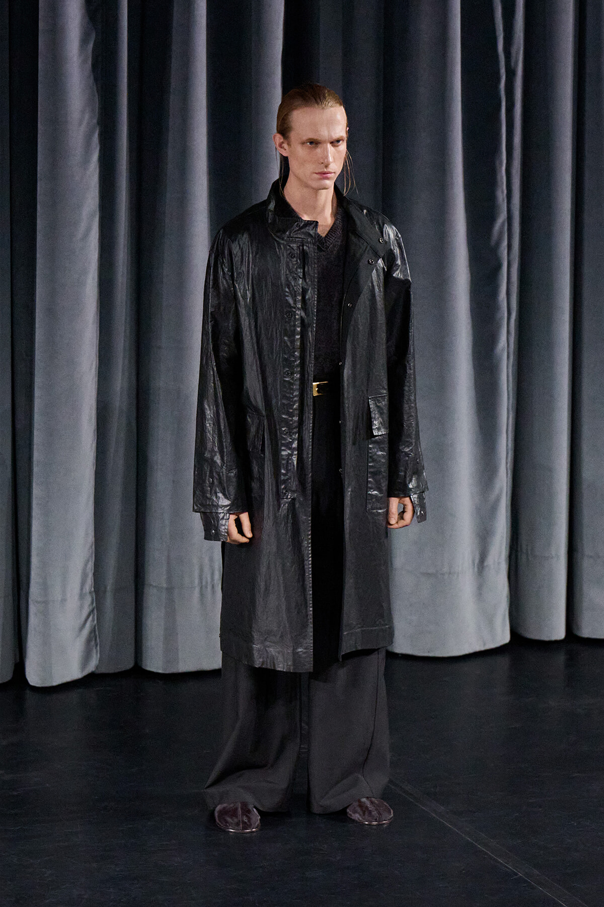 Male model wearing a long black raincoat on a dark stage with gray curtains in the background.