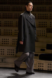 Male model in a charcoal double-breasted overcoat walks on a stage, wearing a black turtleneck and gray wide-leg pants.