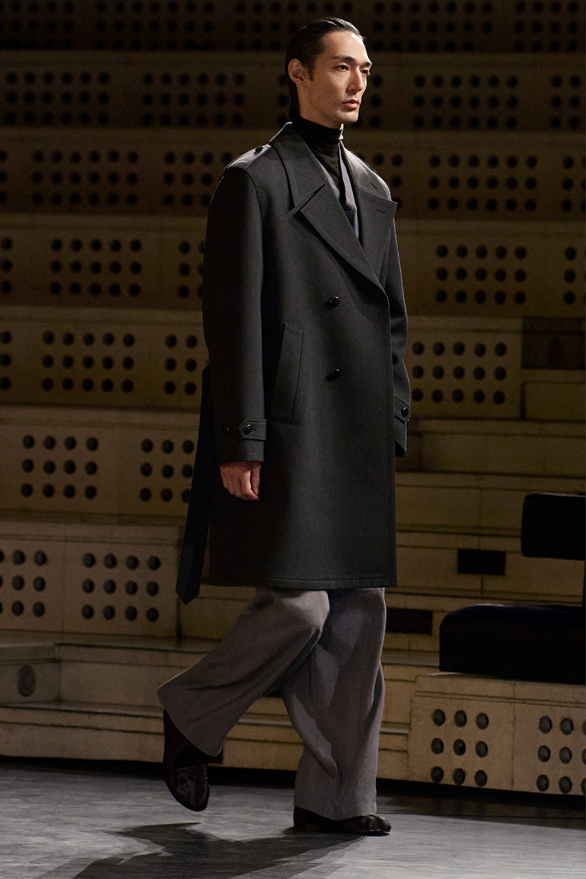 Male model in a charcoal double-breasted overcoat walks on a stage, wearing a black turtleneck and gray wide-leg pants.
