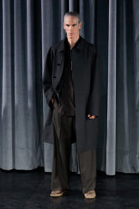 Tall man with gray hair in a black coat stands on a stage in front of dark curtains.