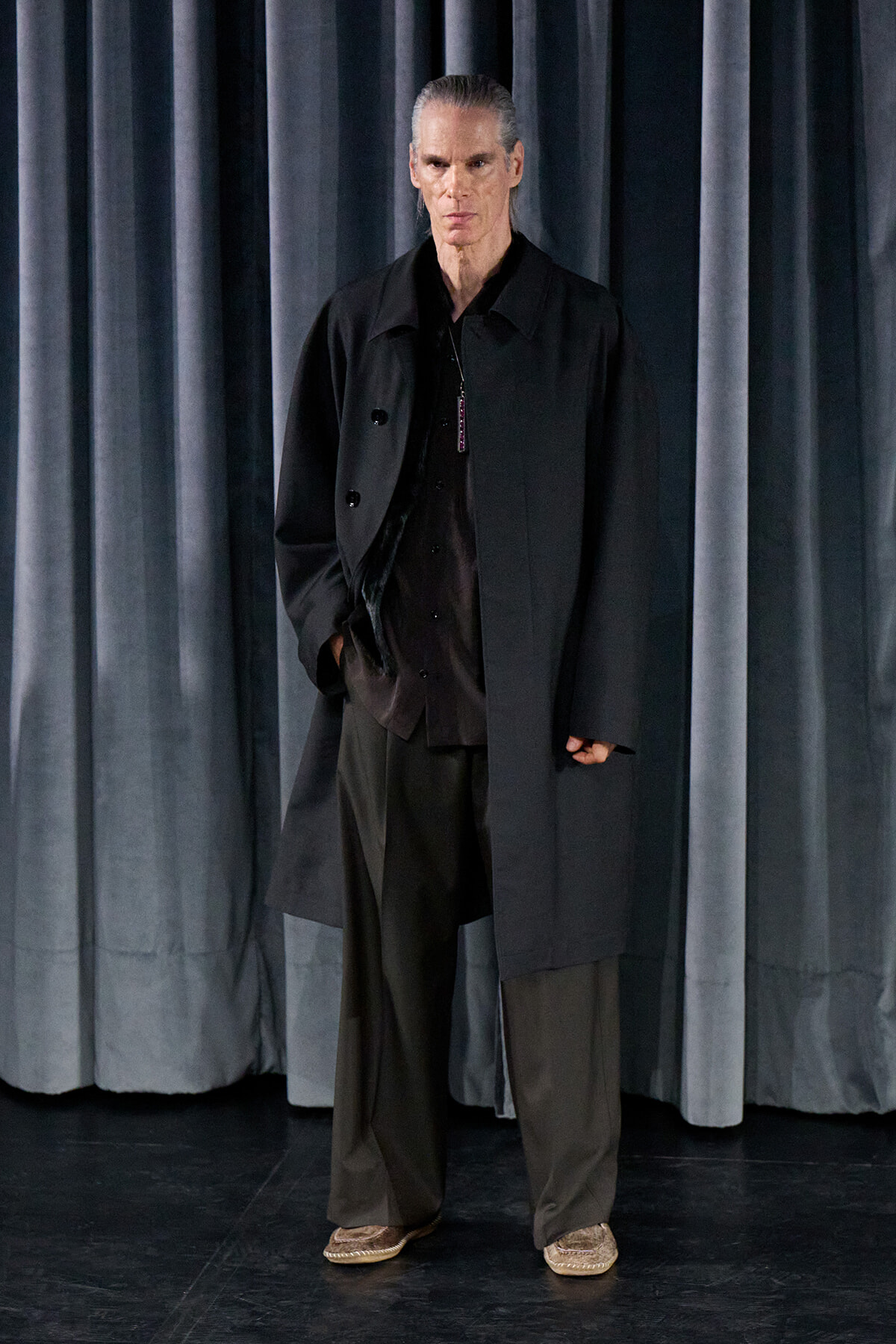 Tall man with gray hair in a black coat stands on a stage in front of dark curtains.