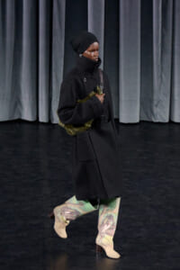 Model in a black high-collar coat and turban walks on a dark stage, holding a green fur bag.