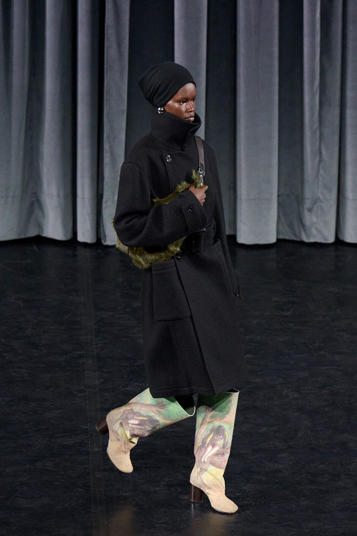 Model in a black high-collar coat and turban walks on a dark stage, holding a green fur bag.