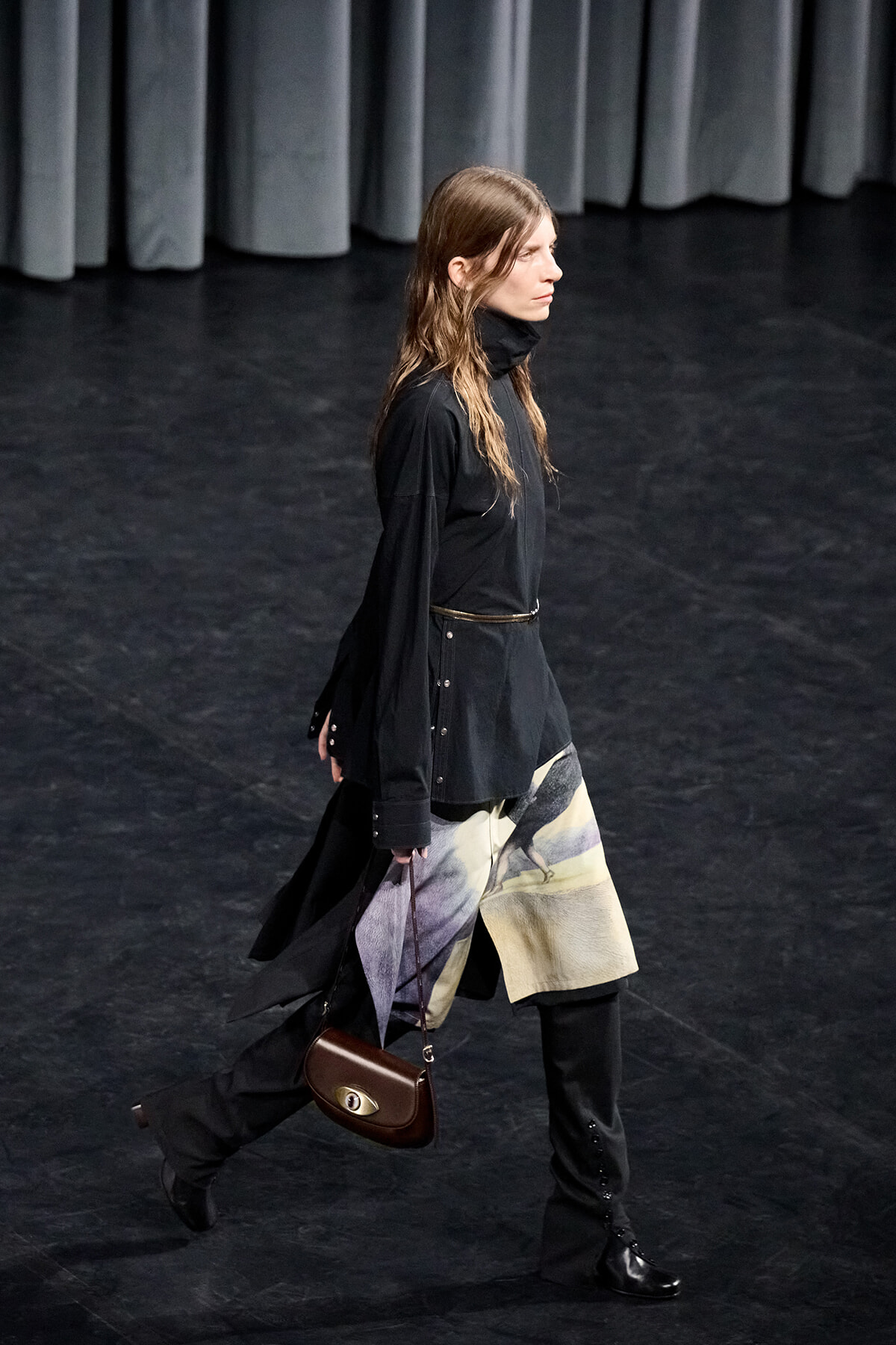 Person walking on a dark stage in a black turtleneck and long black top, paired with a patterned skirt and a small brown handbag.