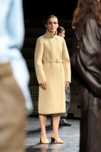 Model wearing a cream knee-length belted wool coat with a high collar on a fashion runway, beige heels visible.