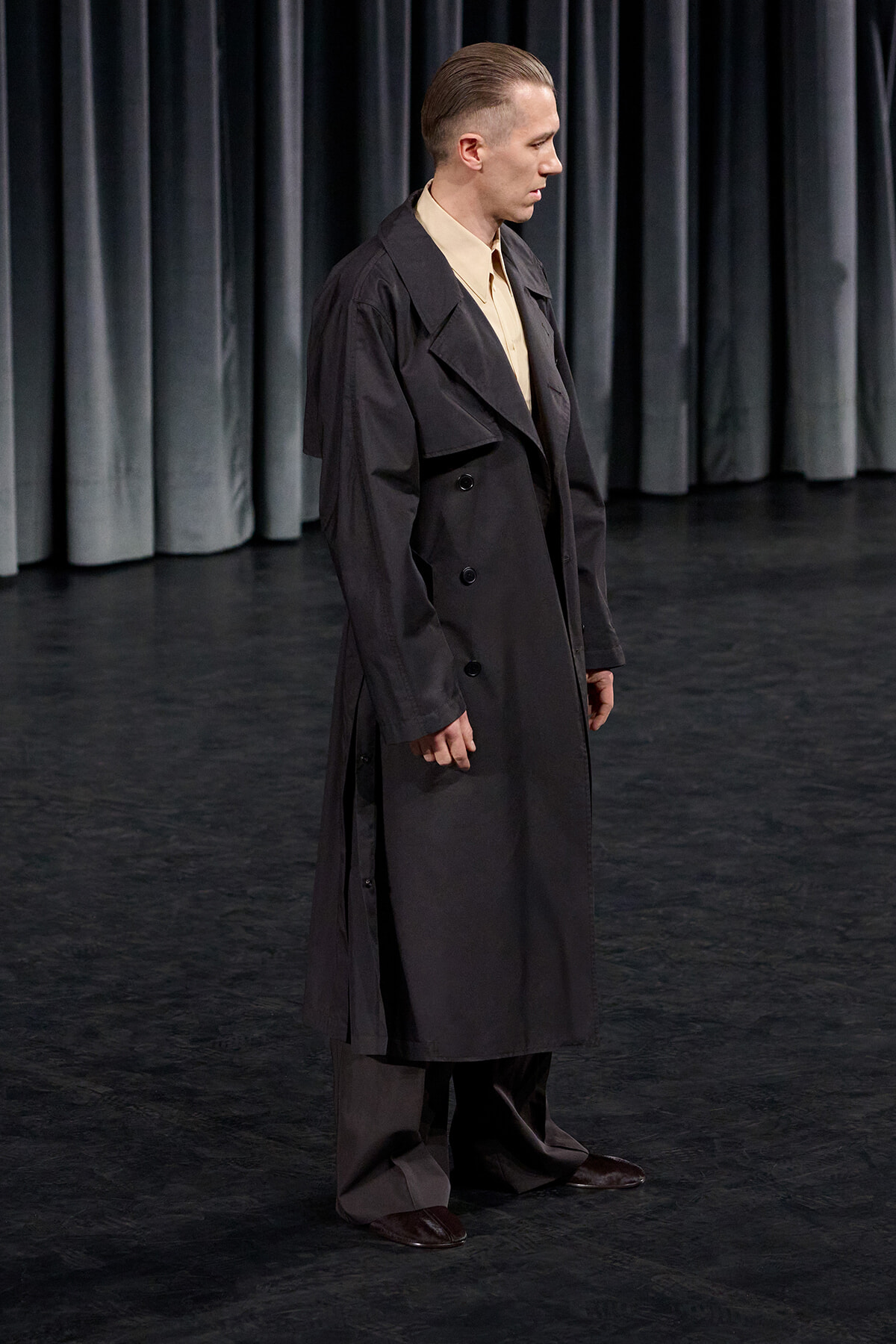 Man in a long black trench coat standing on a stage with dark curtains behind him.