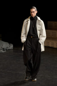 Male model walking a runway in a beige trench over a black turtleneck and oversized black trousers, wearing glasses and a pendant necklace with hands in pockets.