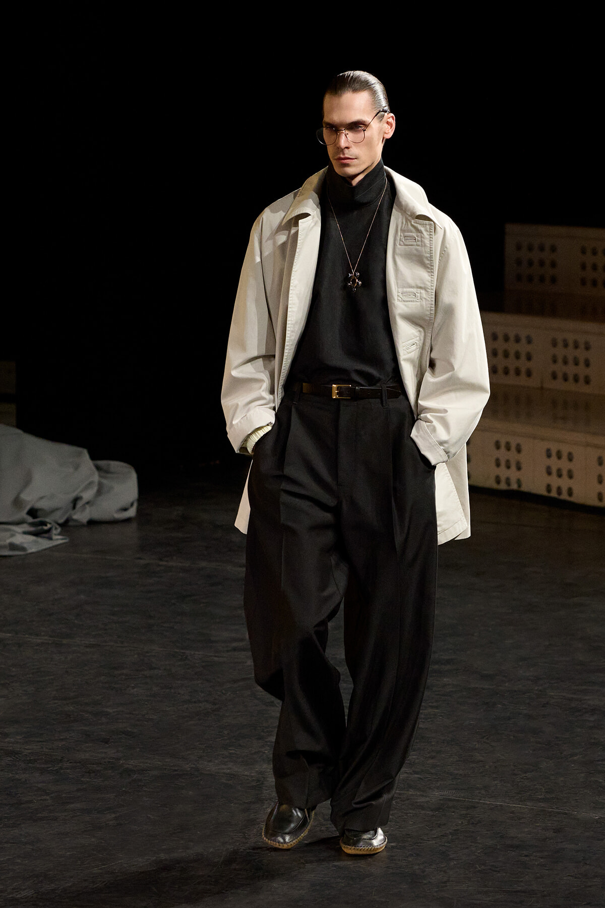Male model walking a runway in a beige trench over a black turtleneck and oversized black trousers, wearing glasses and a pendant necklace with hands in pockets.