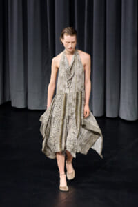 Model walks on a dark stage wearing a flowing, patterned dress with leopard and chevron prints and a draped neckline.
