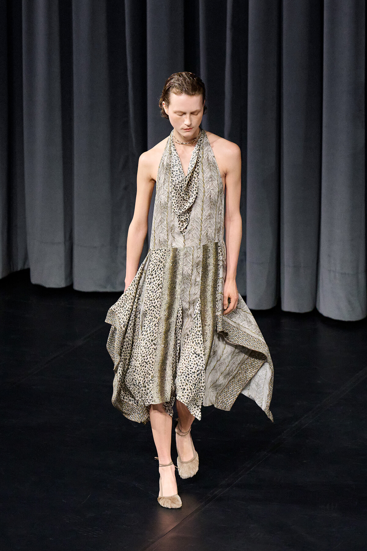 Model walks on a dark stage wearing a flowing, patterned dress with leopard and chevron prints and a draped neckline.