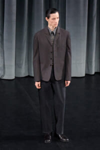 Male model on a dark stage wearing a charcoal blazer, patterned shirt, and black trousers with dress shoes at a fashion show backstage.