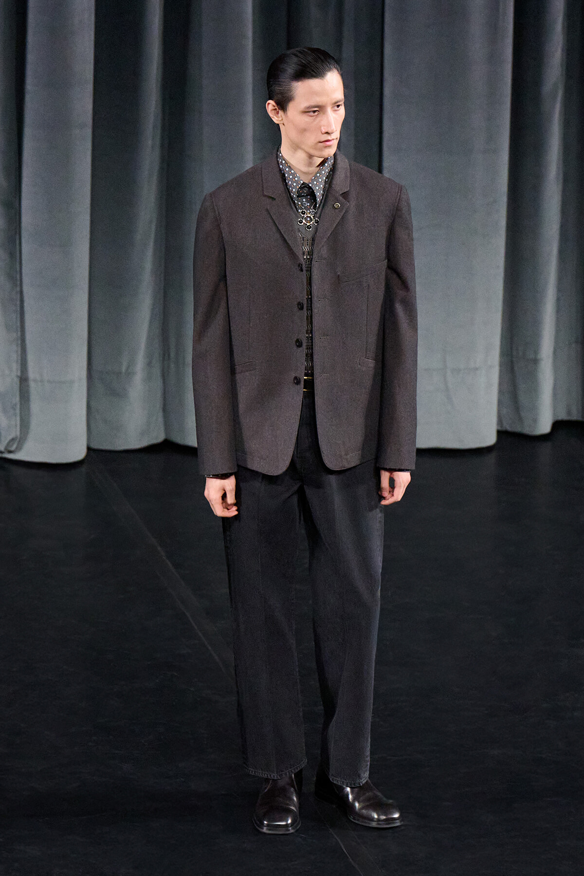 Male model on a dark stage wearing a charcoal blazer, patterned shirt, and black trousers with dress shoes at a fashion show backstage.