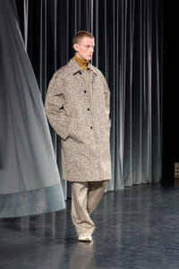 Male model walks on a runway wearing a beige textured overcoat, turtleneck, and loose gray trousers, hands in pockets.