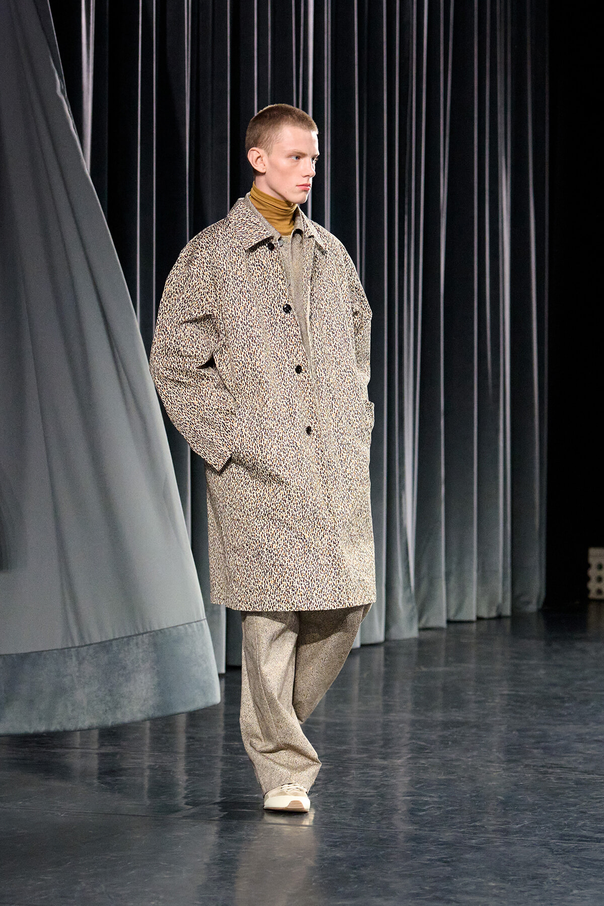 Male model walks on a runway wearing a beige textured overcoat, turtleneck, and loose gray trousers, hands in pockets.