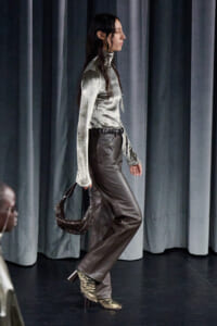 Side-profile of a model walking a runway in a metallic silver turtleneck, brown leather pants, gold heeled boots, and a studded brown handbag.