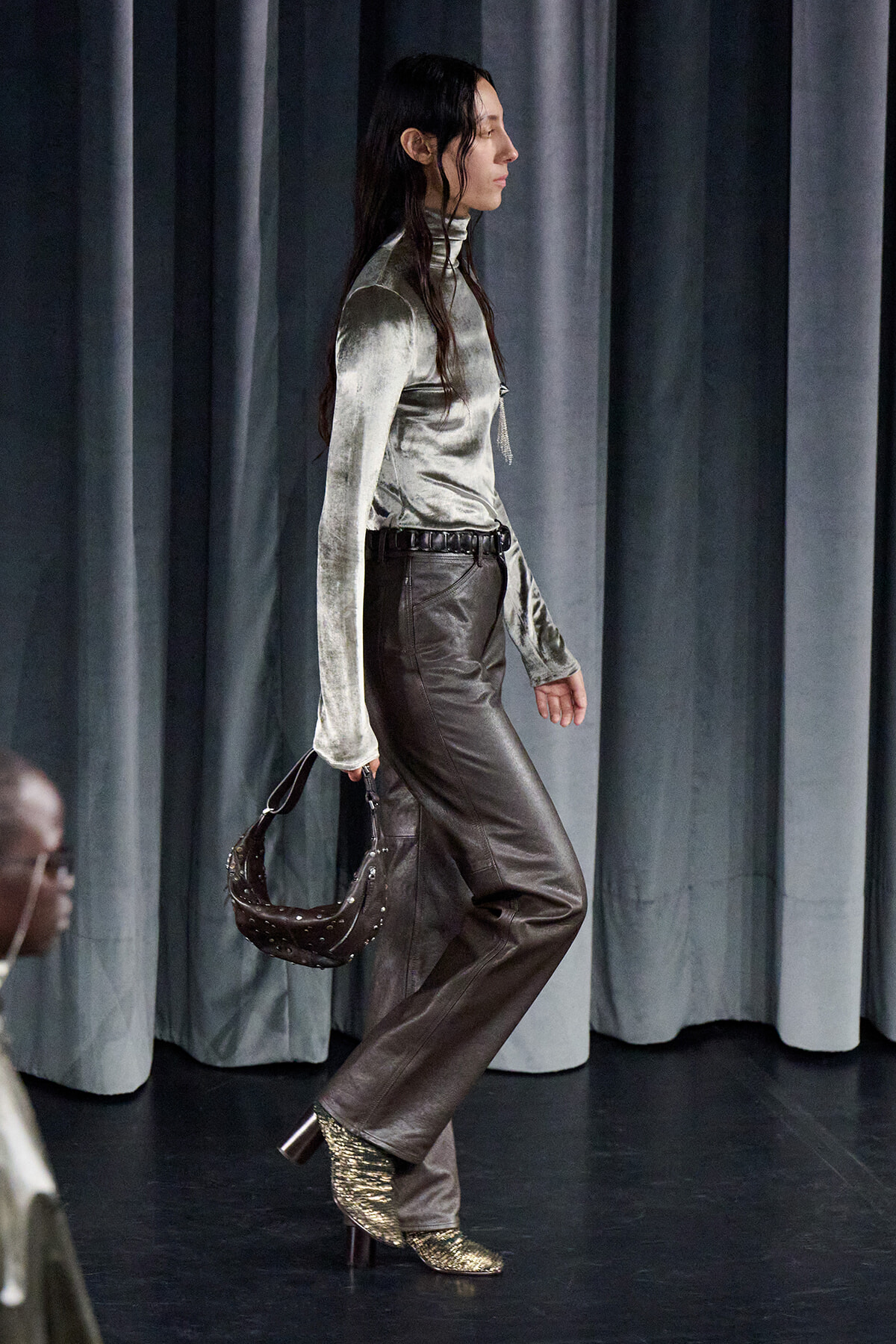 Side-profile of a model walking a runway in a metallic silver turtleneck, brown leather pants, gold heeled boots, and a studded brown handbag.