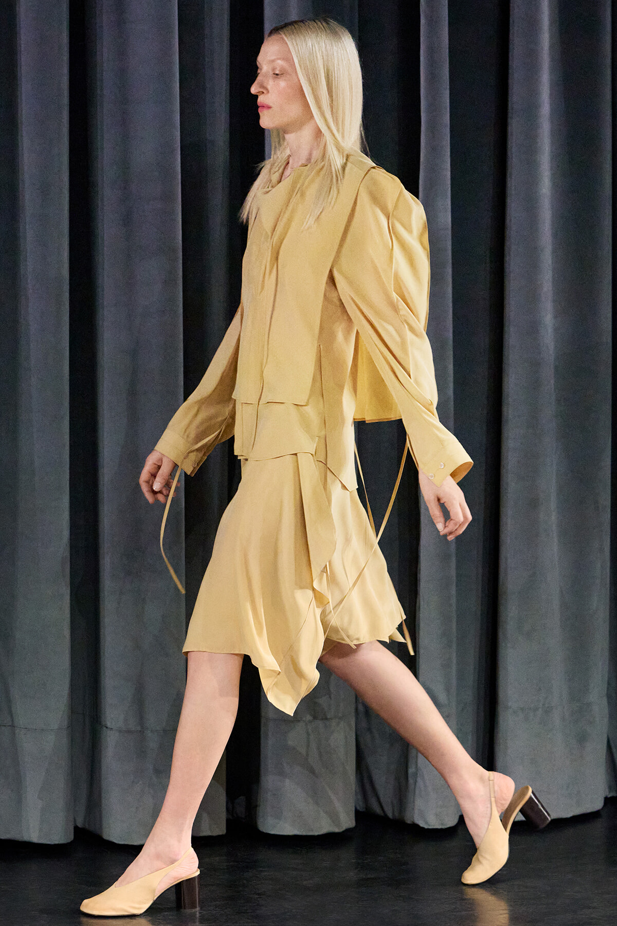 Model walks a beige layered ensemble—a loose trench-style top with draped sleeves and a matching skirt—on a dark stage runway.