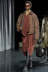 Model on a runway wearing a brown oversized bomber jacket over a rust dress and gold turtleneck, with knee-high boots and sunglasses.