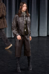 Model on a dark stage wearing a metallic gray leather jacket and matching wide-leg pants with knee-high boots.