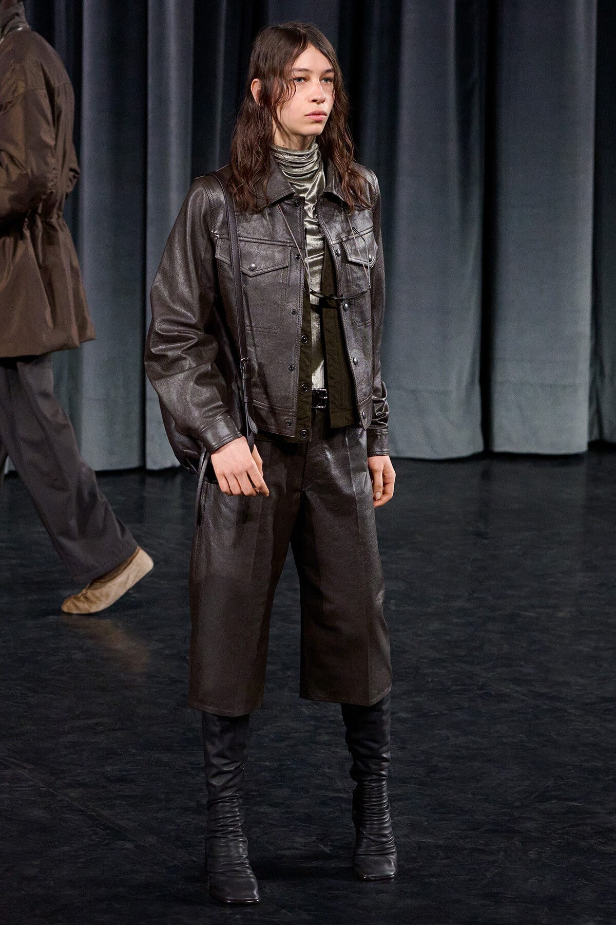 Model on a dark stage wearing a metallic gray leather jacket and matching wide-leg pants with knee-high boots.