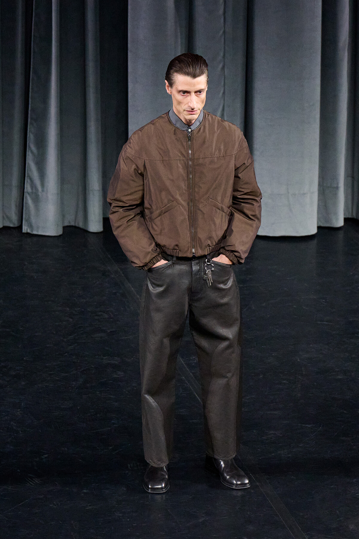 Man with slicked-back hair wearing a brown bomber jacket, gray shirt, and black leather pants stands with hands in pockets on a dark stage with curtains behind him.