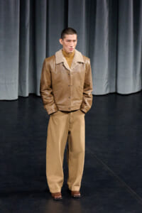 Male model stands on a dark stage wearing a tan shearling-collar leather jacket, camel turtleneck, and khaki wide-leg pants with brown shoes.