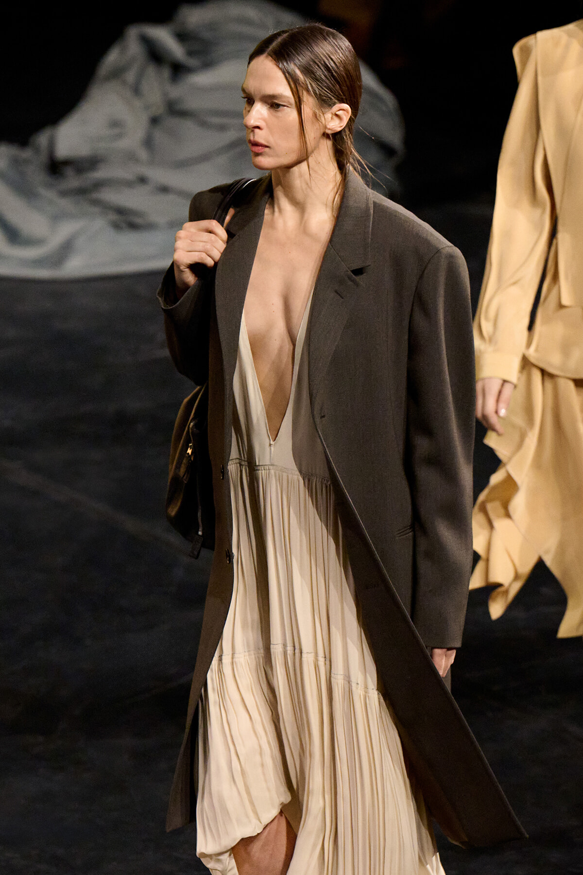 Model walking the runway in a long dark brown coat over a deep V beige pleated dress, carrying a shoulder bag.