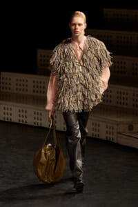 Runway model walking in a shaggy fringe vest over a pink shirt, paired with leather pants and a large brown tote bag