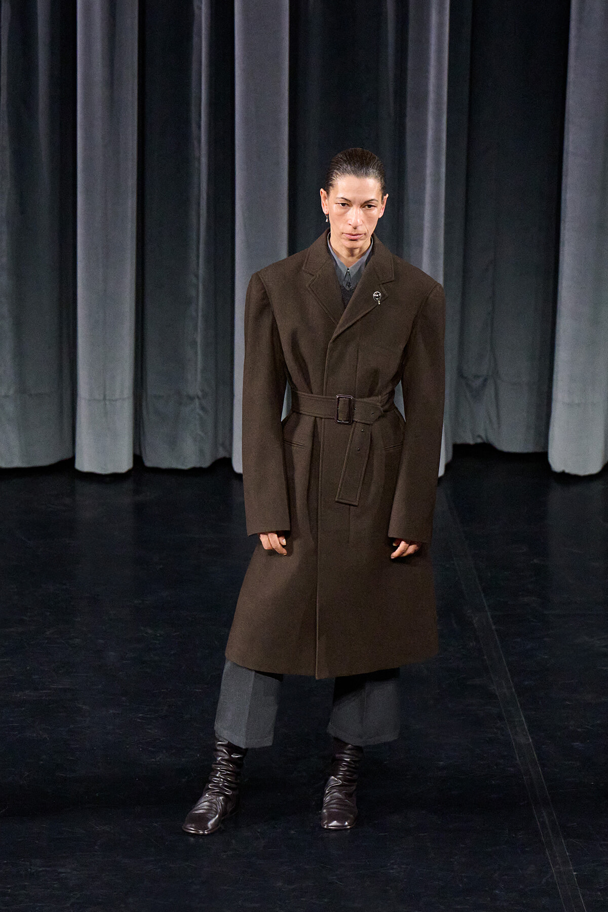 Model stands on a dark stage in a long brown belted trench coat, gray trousers, and brown lace-up boots against gray curtains.